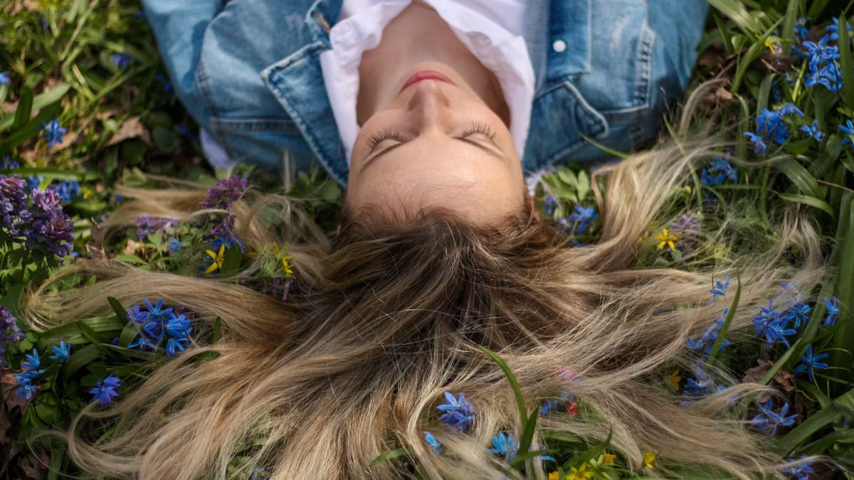 A woman in a denim jacket lies on a lush field of blue and yellow flowers, embracing nature.